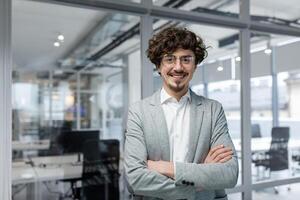 Portrait of successful mature boss, senior businessman in glasses and business suit looking at camera and smiling, man with crossed arms working inside modern office building photo