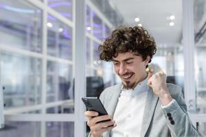 Businessman celebrating success and triumph, man in shirt working inside modern office with laptop holding phone reading message, business owner received good news notification online. photo