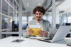 Mature businessman happy with good news received letter in envelope, senior man with beard reading and smiling working inside office at work using laptop, investor celebrating achievement victory. photo