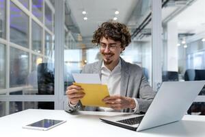 Successful businessman inside office at work, man received notification mail with good news, hispanic man in business suit using laptop at work smiling and reading notification. photo