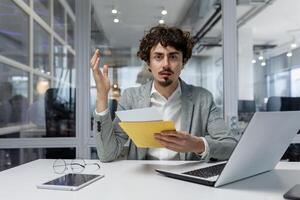 Upset businessman at workplace received letter with bad news, man looking at camera thinking and depressed, worker inside office with laptop. photo