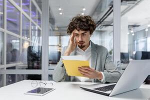 Sad and upset mature businessman received an envelope by mail with a bad news notification letter, boss in business suit working inside office using laptop wearing glasses and beard. photo