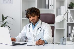 Serious focused hispanic doctor working inside medical office, man in medical coat thinking looking at laptop. photo