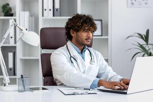 Concentrated young hispanic man doctor student, intern working, studying in hospital in office using laptop. photo