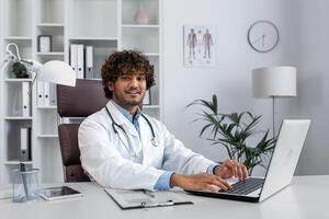 Portrait of a young hispanic doctor working in the office with a laptop, sitting at the table in a white coat and with a stethoscope, smiling and looking at the camera. photo
