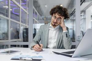 Serious and focused financier accountant on paper work inside office, mature man using calculator and laptop for calculating reports and summarizing accounts, businessman at work in casual clothes. photo