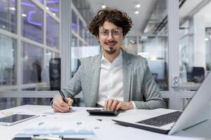 retrato de exitoso empresario detrás papel trabajar, hombre en camisa sonriente y mirando a cámara, financiero jefe dentro oficina utilizando ordenador portátil en trabajo a lugar de trabajo foto