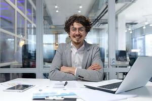 Portrait of successful financier businessman, hispanic man sitting at workplace working with documents and laptop inside office, man with crossed arms smiling and looking at camera. photo