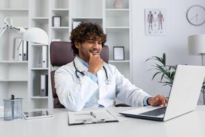 An Indian smiling young doctor in a white coat sits in the office at a table and works on a laptop, talks on the phone with a patient, consults. photo