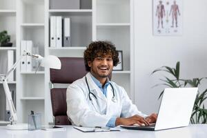 Portrait of young hispanic male medical doctor wearing white coat sitting at desk with laptop in office and smiling at camera. photo