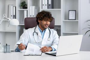 un joven latín americano médico consulta pacientes en línea. él se sienta en el oficina en frente de un ordenador portátil y negociaciones mediante un llamar, escribe abajo el médico historia, hace un encuesta, empieza tratamiento. foto