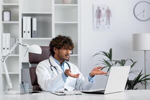 Young indian displeased and serious doctor inside office workplace talking remotely consulting patients using laptop for remote meeting. photo