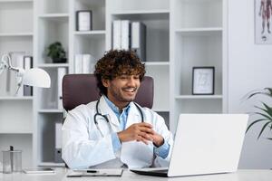 Young successful doctor working inside hospital office, man smiling and looking at laptop screen, hispanic man in medical gown is satisfied with patient's treatment achievement results. photo