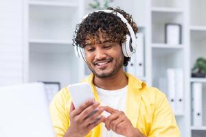 Young smiling hispanic man at home close-up, man in headphones listening to music online books and audio podcasts, using phone typing messages and watching videos, holding smartphone. photo