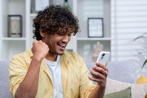 Joyful and happy young man sitting on sofa alone at home, hispanic celebrating victory and success, got good news using app on phone. photo