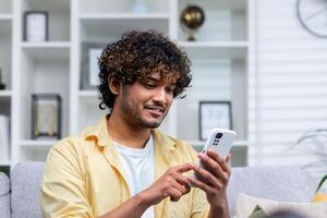 un joven Hispano hombre es sentado a hogar en el sofá en el vivo habitación, el hombre es participación un teléfono en su manos, utilizando un en línea solicitud en un teléfono inteligente, hojeada el Internet y mecanografía mensajes foto