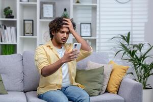 latín americano hombre sentado en sofá a hogar en vivo habitación, hombre trastornado y Deprimido leyendo malo Noticias en línea utilizando aplicación en teléfono, participación teléfono inteligente foto