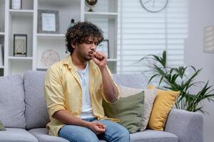 Sick cold man sitting alone on sofa at home, hispanic has bronchitis coughs shortness of breath. photo