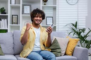 Portrait of young successful man at home on sofa, winner smiling and looking at camera holding phone in hands using game app, holding hand up successful gesture of triumph. photo