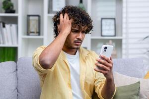 latín americano hombre sentado en sofá a hogar en vivo habitación, hombre trastornado y Deprimido leyendo malo Noticias en línea utilizando aplicación en teléfono, participación teléfono inteligente foto