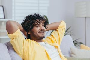 Young man at home on the sofa dreaming smiling and looking out the window, Indian man with his hands behind his head in the living room thinking with a smile. photo