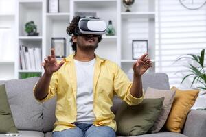 Latin American man sitting on couch at home using virtual reality glasses, man viewing educational material remotely. photo