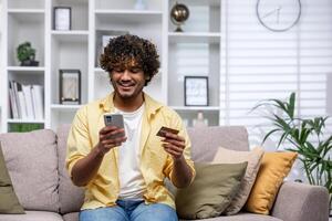 joven Hispano hombre a hogar solo en sofá en vivo habitación sentado sonriente y feliz, hombre utilizando teléfono y banco crédito tarjeta a tienda en línea en en línea almacenar. foto