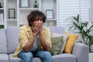 Sad depressed man sitting alone at home on sofa in living room, desperate indian man. photo