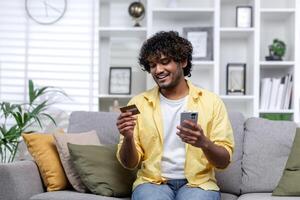 joven Hispano hombre a hogar solo en sofá en vivo habitación sentado sonriente y feliz, hombre utilizando teléfono y banco crédito tarjeta a tienda en línea en en línea almacenar. foto