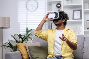 Latin American man sitting on couch at home using virtual reality glasses, man viewing educational material remotely. photo
