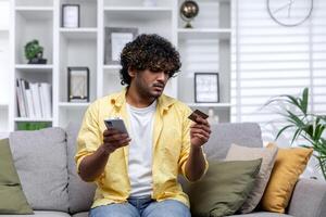 Cheated man at home trying to make online purchase and money transfer sitting on sofa in living room, Hindu man holding bank credit card and using online payment app on phone. photo