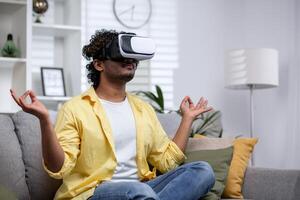 Young Hindu man in yellow shirt alone at home sitting on sofa and meditating in lotus position, man using vr glasses for virtual meditation and travel. photo