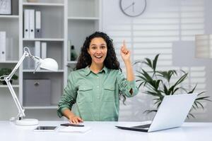 A young muslim woman sits in the office at a desk with a laptop, looks at the camera. points his finger up, indicates, has an idea, a plan. photo