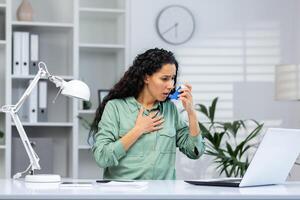 Asthma attack in Hispanic business woman inside office at workplace with laptop, woman uses inhaler with medication to ease breathing. photo