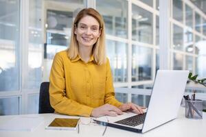 mujer de negocios alegre a lugar de trabajo utilizando teléfono, latín americano contento utilizando aplicación en teléfono inteligente, hojeada social redes, trabajando en hogar oficina sentado con ordenador portátil. foto