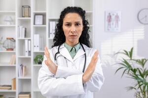 Close-up portrait of young serious hispanic female doctor standing in hospital office in white coat and crossed arms showing no sign to camera. photo