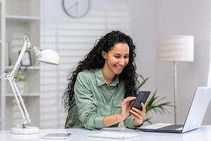 mujer de negocios alegre a lugar de trabajo utilizando teléfono, latín americano contento utilizando aplicación en teléfono inteligente, hojeada social redes, trabajando en hogar oficina sentado con ordenador portátil. foto