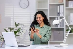 mujer de negocios alegre a lugar de trabajo utilizando teléfono, latín americano contento utilizando aplicación en teléfono inteligente, hojeada social redes, trabajando en hogar oficina sentado con ordenador portátil. foto