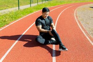 Sports injury. A young Asian sportsman, athlete, runner sits on a stadium treadmill and holds his leg, knee. feels severe pain. photo