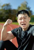 Close-up vertical photo. Young Asian man sitting in the stadium after playing sports in headphones and groping for the bell, showing a hand gesture of strength and victory. photo