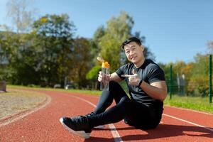 Young Asian smiling man resting after running and doing sports. Sitting on the track of the stadium, holding water in his hands and pointing to the bottle. photo