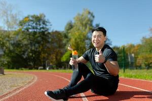Tired and smiling young asian male runner sitting and resting on stadium track, holding water bottle and showing super gesture with finger. photo