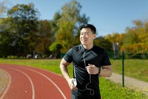 Young Asian male athlete running in the stadium, doing a morning run in headphones, holding a phone, listening to music. audiobook, podcast. photo