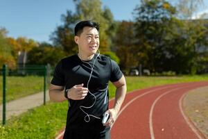 Asian smiling young man doing sports and running in stadium wearing headphones, holding phone. photo