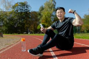 Portrait of a young Asian man, an athlete sitting on a treadmill with a phone. He is happy, looks into the camera. Shows a gesture of victory and strength. Checks the results of running, training. photo