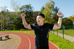 asiático joven masculino corredor y deportista en pie en estadio, levantamiento manos arriba, alegría y celebrando victoria a terminar línea, satisfecho con resultado. foto