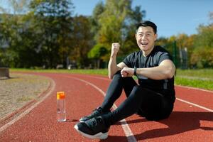 A young Asian man, an athlete sitting on a stadium treadmill. He looks at the smartwatch, fitness bracelet. Shows a gesture of victory and strength. Checks the results of running, training. photo