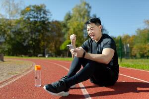 Portrait of a young Asian man, an athlete sitting on a treadmill with a phone. Rejoicing, showing a victory gesture, yes. Checks the results of jogging, training, celebrates. photo