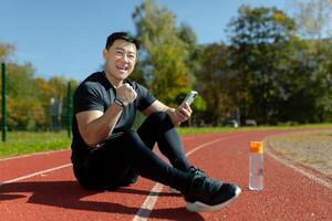 Joyful and successful man resting after fitness and active physical exercises, looking at camera and celebrating good results, using app on smartphone for training. photo