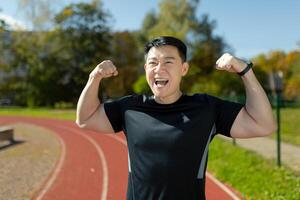 victorioso el competencia. un joven asiático hombre, un atleta, un corredor celebra un victoria en un carrera. contento hombre en pie en estadio en corriendo pista y demostración fuerza y victoria gestos con su manos. foto